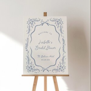 May include: A white sign on a wooden easel with blue floral and ribbon accents. The sign reads "Welcome to Isabelle's Bridal Shower" and includes the date and location, "The Baltimore Hotel."