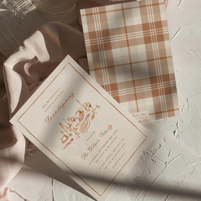 K&ouml;nnte beinhalten: Zwei cremefarbene Einladungen mit braunem Text und Illustrationen. Eine zeigt den Text "Friendsgiving" und Veranstaltungsdetails. Die andere hat ein braunes und wei&szlig;es Karomuster. Die Einladungen liegen auf einer wei&szlig;en Oberfl&auml;che mit einem weichen rosa Stoff.