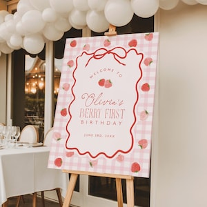 May include: A pink and white gingham welcome sign on a wooden easel. The sign has a red border and features the text "Welcome to Olivia's Berry First Birthday" with strawberry illustrations. White balloons are visible in the background.