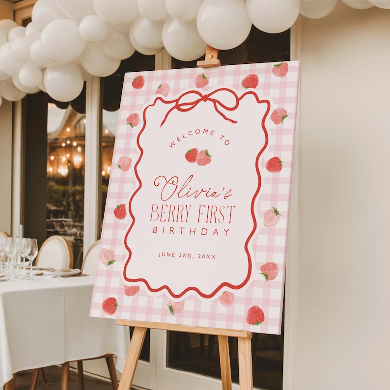 May include: A pink and white gingham welcome sign on a wooden easel. The sign has a red border and features the text "Welcome to Olivia's Berry First Birthday" with strawberry illustrations. White balloons are visible in the background.