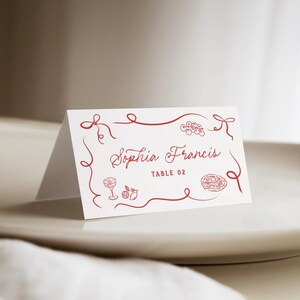 May include: A folded white place card with red decorative border and text. The card reads "Sophia Francis" and "TABLE 02". The design includes illustrations of a cocktail, fruit, and pizza.
