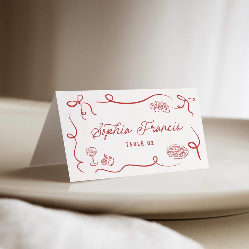 May include: A folded white place card with red decorative border and text. The card reads "Sophia Francis" and "TABLE 02". The design includes illustrations of a cocktail, fruit, and pizza.