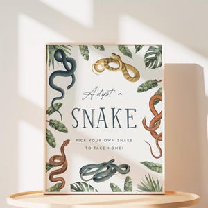May include: A rectangular sign with a white background featuring illustrations of various snakes and green tropical leaves. The text reads: Adopt a SNAKE. PICK YOUR OWN SNAKE TO TAKE HOME! The snakes are depicted in shades of blue, brown, and gold.