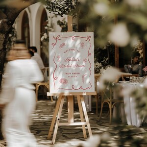 May include: A white sign on a wooden easel with red text and illustrations. The sign reads "Welcome to Sophia's Bridal Shower" with the date "04.25.XX" and the phrase "Thats Amore!" The background is a blurred outdoor setting.