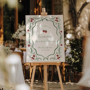 May include: A welcome sign on a wooden easel. The sign has a white background with a green border and red berry accents. The text reads "Welcome to Isabelles Bridal Shower" and "The Elm's Garden".