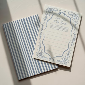 Op de afbeelding: Twee notitieboekjes, één met blauw-witte strepen en de andere met de tekst "Love Letter to the Bride" in blauw schrift. Het briefpapier heeft een bloemenrand en lijnen om op te schrijven. De tekst luidt: "Schrijf een oprechte brief aan de bruid met je beste wensen, huwelijksadvies of een gekoesterde herinnering. Dit wordt een aandenken voor altijd!"