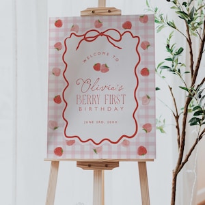 May include: A wooden easel displays a sign with a pink gingham background and a decorative red border. The sign reads "Welcome to Olivia's Berry First Birthday" with strawberry illustrations. The date "June 3rd, 20XX" is also included.