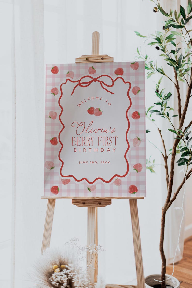 May include: A wooden easel displays a sign with a pink gingham background and a decorative red border. The sign reads "Welcome to Olivia's Berry First Birthday" with strawberry illustrations. The date "June 3rd, 20XX" is also included.