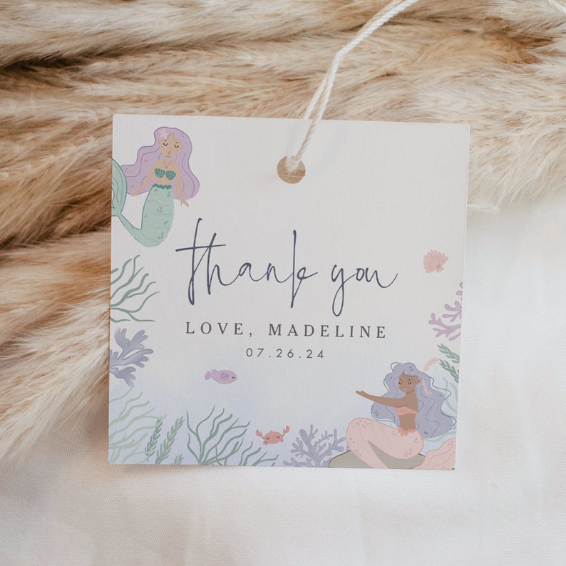 Puede incluir: Una etiqueta de agradecimiento con una ilustraci&oacute;n de sirena. La etiqueta es blanca con un fondo azul claro y presenta una sirena morada y verde con una cola rosa. El texto en la etiqueta dice "gracias" en cursiva y "LOVE, MADELINE 07.26.24".