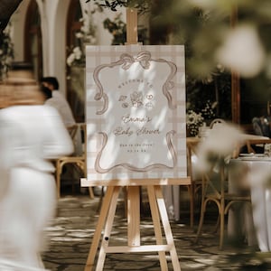 May include: A wooden easel displays a baby shower sign. The sign features a beige and white checkered background with a decorative border and the words "Welcome to Emma's Baby Shower." The sign also includes illustrations of baked goods.