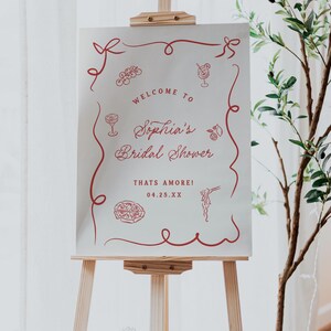 May include: A wooden easel displays a white sign with red text and illustrations. The sign reads "Welcome to Sophia's Bridal Shower" with decorative elements. The date is 04.25.XX. A plant is to the right.