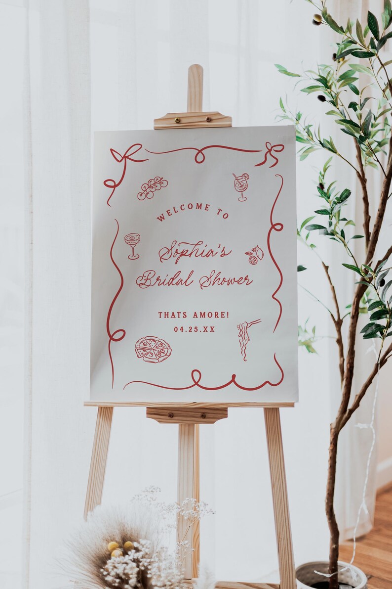 May include: A wooden easel displays a white sign with red text and illustrations. The sign reads "Welcome to Sophia's Bridal Shower" with decorative elements. The date is 04.25.XX. A plant is to the right.
