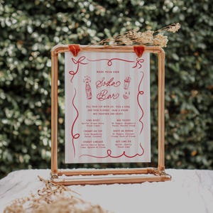 May include: A copper-colored metal frame holds a sign for a soda bar. The sign has a red and white striped background with red text and illustrations. The text reads "Make Your Own Soda Bar" and lists soda flavor combinations.