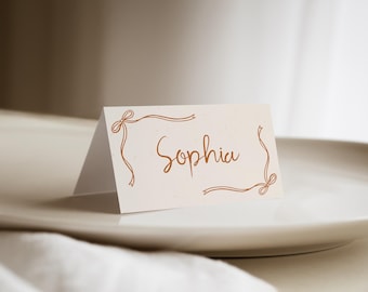 May include: A folded white place card featuring the name Sophia written in a brown cursive script, framed by a delicate brown line drawing of ribbons and bows at the top left and bottom right corners on a textured paper surface.