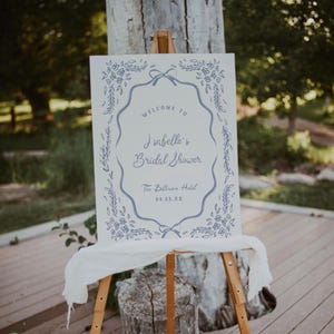 May include: A white sign on a wooden easel with blue floral and bow accents. The sign reads "Welcome to Isabelle's Bridal Shower" and includes the date and location, The Biltmore Hotel.