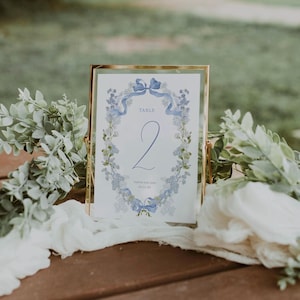 May include: A white table number card with a blue floral wreath and a blue bow. The card says "Table 2" and "Sophia and John 06.17.23".