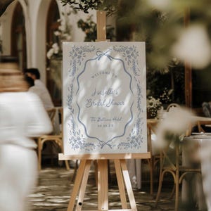 May include: A white sign on a wooden easel with blue floral and ribbon accents. The sign reads "Welcome to Isabelle's Bridal Shower" and includes the date and location.