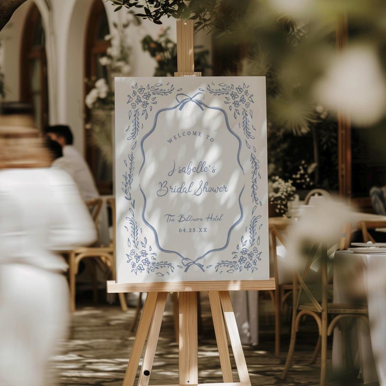 May include: A white sign on a wooden easel with blue floral and ribbon accents. The sign reads "Welcome to Isabelle's Bridal Shower" and includes the date and location.