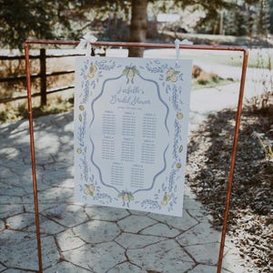 May include: A seating chart for Isabelle's Bridal Shower, displayed on a copper frame. The white paper features a blue border with lemon and leaf illustrations. The text includes table numbers and space for guest names.