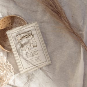 May include: A rectangular invitation card with a beige and white striped border featuring a black line drawing of a storefront with an awning and the text Chloe's 1st Birthday, A brew is brewing, Saturday, June 25, 2 PM, 5 Sapphire Street, Any City, RSVP to 555-0100.