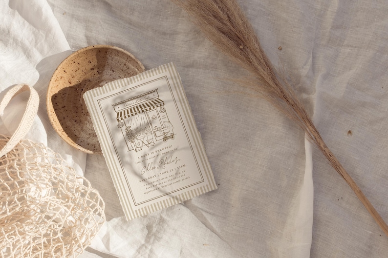 May include: A rectangular invitation card with a beige and white striped border featuring a black line drawing of a storefront with an awning and the text Chloe's 1st Birthday, A brew is brewing, Saturday, June 25, 2 PM, 5 Sapphire Street, Any City, RSVP to 555-0100.