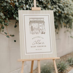 Op de afbeelding: Een rechthoekig welkomstbord voor een babyshower met een beige en wit verticaal gestreepte rand. Het midden toont een lijntekening van een cafégevel met een kinderwagen en de tekst: Welcome to Chloe's Baby Shower 03.15.xx.