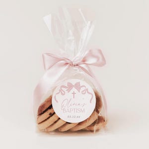 May include: A clear cellophane bag tied with a pink ribbon, containing several cookies. A round sticker with a pink bow, cross, and the text "Olivia's Baptism" is on the front. The cookies are light brown with dark chocolate chips.