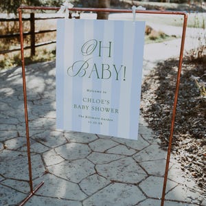 Pu&ograve; includere: Una cornice in metallo color rame sostiene un cartello con la scritta "OH BABY!" in verde. Il cartello ha uno sfondo a righe blu e bianche e d&agrave; il benvenuto agli ospiti del baby shower di Chloe al Biltmore Garden.