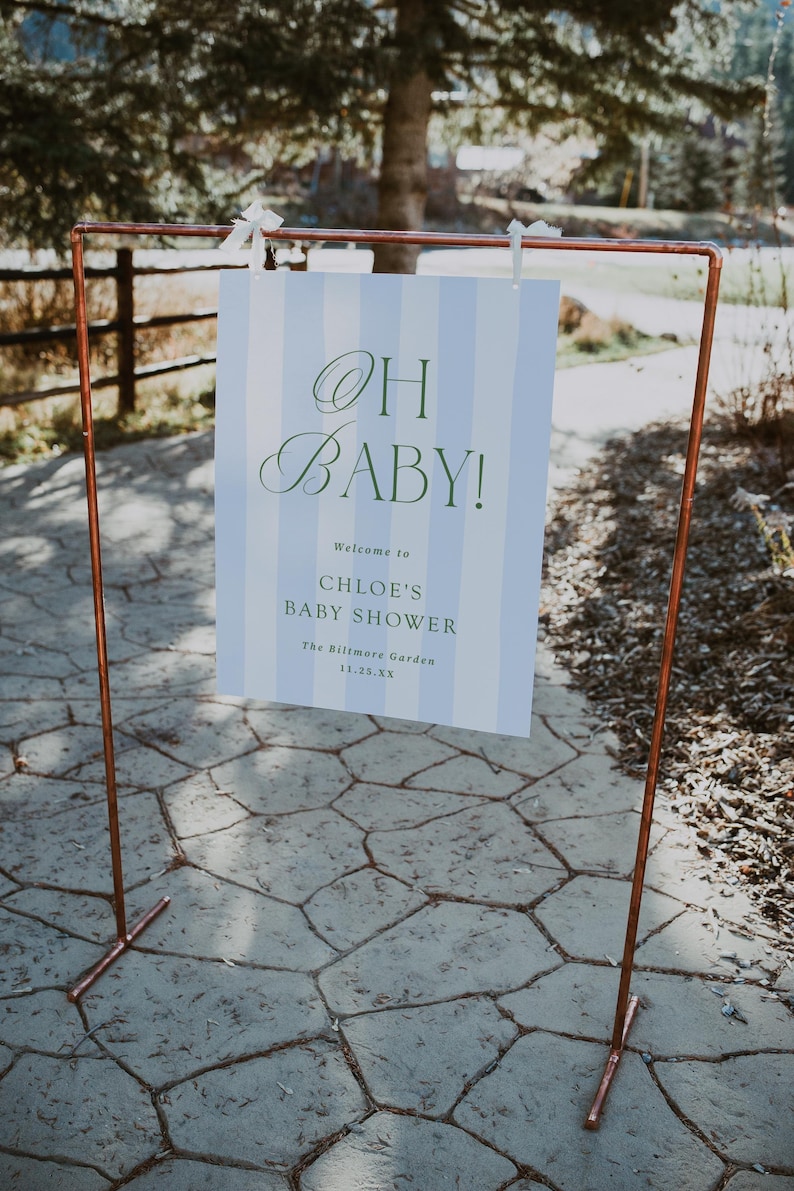 Pu&ograve; includere: Una cornice in metallo color rame sostiene un cartello con la scritta "OH BABY!" in verde. Il cartello ha uno sfondo a righe blu e bianche e d&agrave; il benvenuto agli ospiti del baby shower di Chloe al Biltmore Garden.