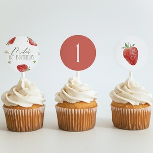 May include: Three cupcakes with white icing and decorative toppers. One topper says "Mila's 1st Birthday" with strawberry illustrations. Another has a red circle with the number "1". The third topper features a strawberry.