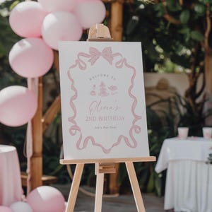May include: A pink and white welcome sign with a pink ribbon bow and the text "Welcome to Olivia's 2nd Birthday Let's Par-Tea!" The sign is on a wooden easel.