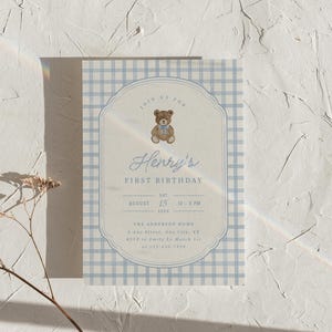 May include: A rectangular birthday invitation card featuring a light blue and white gingham border. The center displays a brown teddy bear with a blue bow, the text Henry's FIRST BIRTHDAY, and event details including date, time, and location.