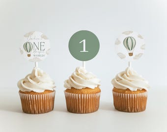 May include: Three cupcakes topped with circular paper decorations on white sticks. The left topper features a hot air balloon and the text Jackson's ONE derful WORLD. The center is a solid sage green circle with the number 1. The right shows a balloon.