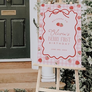 May include: A pink and white gingham sign on a wooden easel. The sign reads "Welcome to Olivia's Berry First Birthday" with a strawberry border and bow. The date is June 3rd, 20XX.