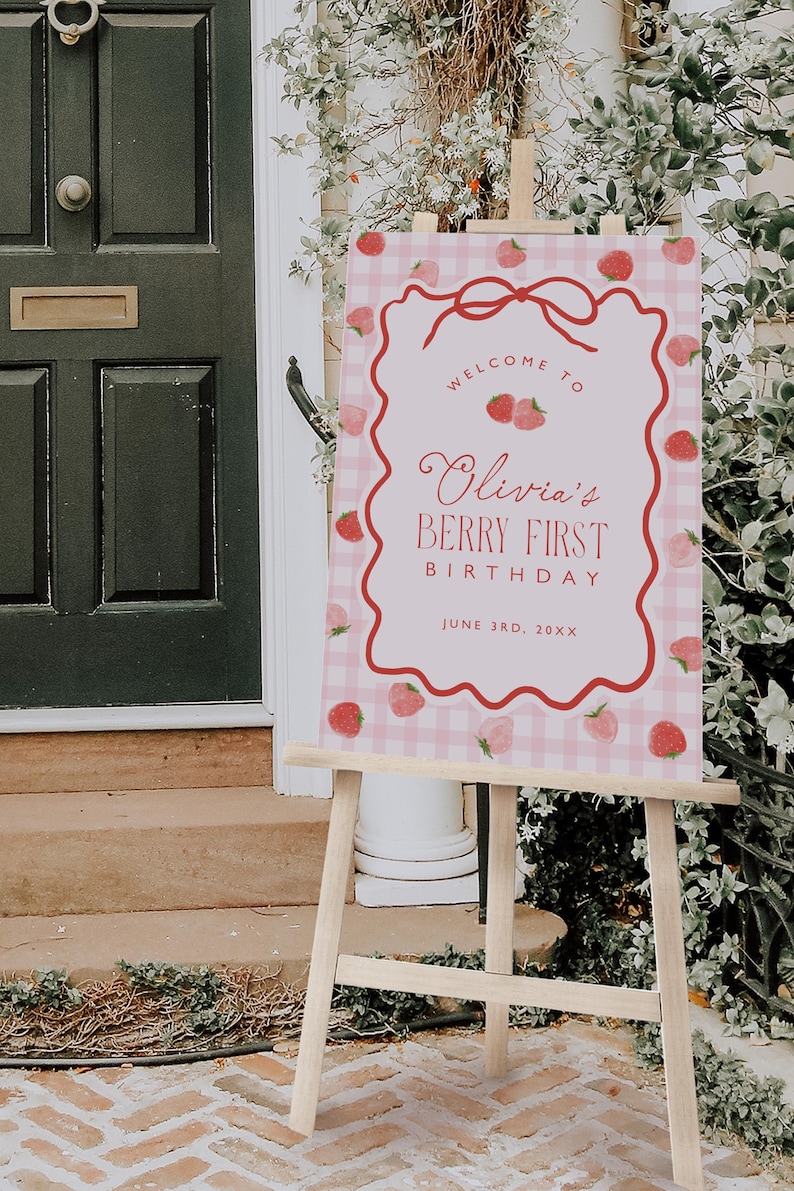 May include: A pink and white gingham sign on a wooden easel. The sign reads "Welcome to Olivia's Berry First Birthday" with a strawberry border and bow. The date is June 3rd, 20XX.