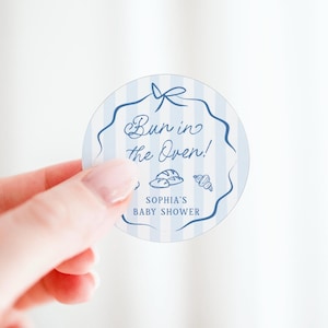 Può includere: Un adesivo rotondo con sfondo a righe blu e bianche e un bordo blu. Mostra il testo "Bun in the Oven!" con illustrazioni di pane e croissant. È presente anche il testo "SOPHIA'S BABY SHOWER".
