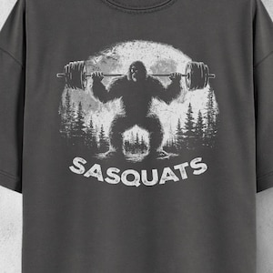 May include: Pepper-colored t-shirt featuring a white graphic of a Bigfoot lifting weights with the text "SASQUATS" below. The design includes a forest and a full moon. The shirt is hanging on a wooden hanger.