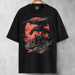 Puede incluir: Camiseta negra con un diseño de un dragón frente a una luna roja y un castillo. El diseño presenta un dragón, un castillo sobre un terreno rocoso y una luna roja. La camiseta está colgada en una percha de madera.