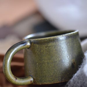 Op de afbeelding: Een groene keramische mok met een afgeronde vorm en een handvat. De mok heeft een getextureerd oppervlak en staat op een bruin en wit doek.