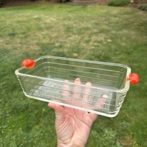 Könnte beinhalten: Eine rechteckige Auflaufform aus klarem Glas mit horizontal gerippten Seiten und zwei leuchtend orangefarbenen Kunststoffgriffen an den Enden. Die Form hat ein tiefes Becken und eine transparente Konstruktion.