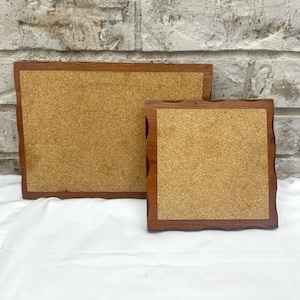 May include: Two wooden coasters with a cork centre. The coasters have a dark brown, wavy edge. One coaster is square, and the other is rectangular. The background is a white surface and a brick wall.