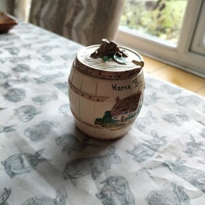 May include: A white ceramic honey pot shaped like a barrel with a cottage scene and the text "Herne Bay" on the side. The lid has a small frog figurine on top.