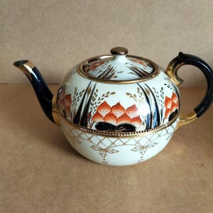 May include: A white teapot with a black handle and spout. The teapot has a gold trim and a floral design in shades of orange, black, and gold.