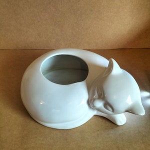 May include: White ceramic planter shaped like a sleeping cat. The planter has a round opening on the top for planting.