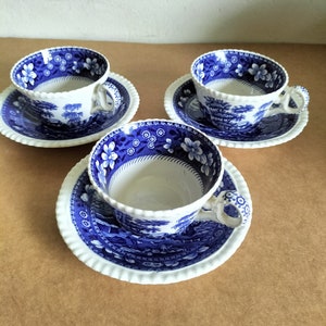 May include: Three white teacups and saucers with a blue floral and landscape pattern. The cups have handles and sit on matching saucers with a scalloped edge. The blue design is on the inside and outside of the cups and saucers.