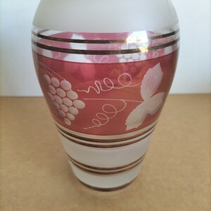 May include: A frosted glass vase with a pink and gold design. The vase has a wide mouth and a narrow base. The design features a grapevine with leaves and grapes.