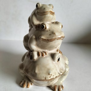 May include: A stack of three ceramic frogs, each with a different expression. The bottom frog is the largest and has a brown glaze, while the top two frogs are smaller and have a white glaze.