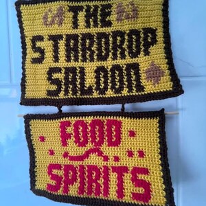 May include: A two-tiered crocheted wall hanging featuring yellow rectangular panels with dark brown borders. The top panel displays the text THE STARDROP SALOON in dark brown, while the bottom panel features the text FOOD SPIRITS in red.