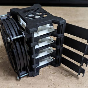 May include: A black 3D-printed rack designed to hold four white single-board computers in a vertical stack. The assembly features a large black cooling fan attached to the front with metal screws and four extended black arms on the side.