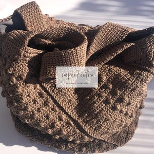 Puede incluir: Un bolso tote de crochet marrón con un diseño texturizado y abultado. El bolso está doblado y tiene una parte superior grande y abierta.
